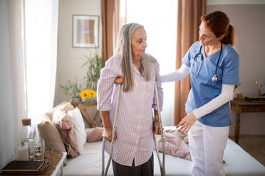 Caregiver helping senior woman to walk with crutches in her home. Thoughful nurse taking care of eldery patient after surgery. - HPIF34435