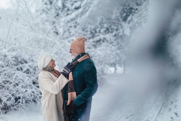 Elegant senior couple walking in the snowy park, during cold winter snowy day. Elderly woman fasten husband's winter coat. Wintry christmas landscape. - HPIF34469