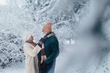 Elegantes älteres Paar, das im verschneiten Park spazieren geht, während eines kalten, verschneiten Wintertages. Ältere Frau befestigt den Wintermantel ihres Mannes. Winterliche Weihnachtslandschaft. - HPIF34469