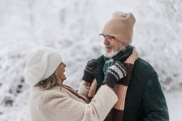 Elegantes älteres Paar, das im verschneiten Park spazieren geht, während eines kalten, verschneiten Wintertages. Ältere Frau befestigt den Wintermantel ihres Mannes. Winterliche Landschaft. - HPIF34470