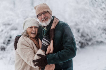 Waist up portrait of elegant senior couple walking in the snowy park, during cold winter snowy day. Elderly couple spending winter vacation in the mountains. Wintry landscape. - HPIF34472