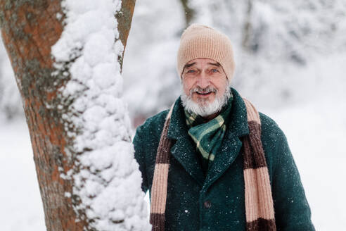 Porträt eines eleganten älteren Mannes, der an einem kalten, verschneiten Wintertag unter einem Baum steht. Älterer Mann, der seinen Winterurlaub in den Bergen verbringt. Winterliche Landschaft. - HPIF34480