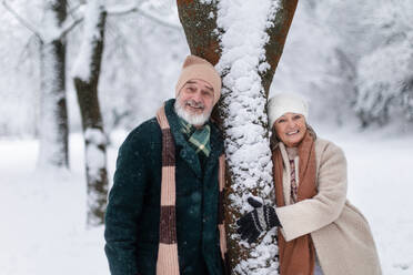 Elegantes älteres Paar, das an einem Baum im verschneiten Park steht, während eines kalten, verschneiten Wintertages. Älteres Paar, das Winterurlaub in den Bergen verbringt. Winterliche Landschaft. - HPIF34482