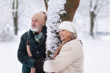 Elegantes älteres Paar, das an einem Baum im verschneiten Park steht, während eines kalten, verschneiten Wintertages. Älteres Paar, das Winterurlaub in den Bergen verbringt. Winterliche Landschaft. - HPIF34485
