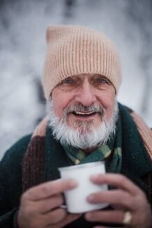 Portrait of elegant senior man drinking hot tea outdoors, during cold winter snowy day. Elderly man spending winter vacation in the mountains. Wintry landscape. - HPIF34509