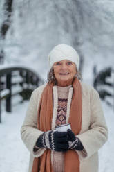 Porträt einer eleganten älteren Frau, die einen heißen Tee im Freien trinkt, während eines kalten, verschneiten Wintertages. Ältere Frau, die Winterurlaub in den Bergen verbringt. Winterliche Weihnachtslandschaft. - HPIF34510