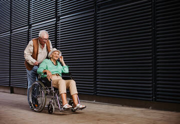 Porträt eines älteren Paares mit Frau im Rollstuhl. Älterer Mann schiebt seine schöne Frau im Rollstuhl. Konzept der chronischen Krankheit bei einem älteren Paar. Porträt mit Kopierraum auf schwarzem Hintergrund. Ältere Geschwister auf einem Sonntagsspaziergang in der Stadt. - HPIF34548