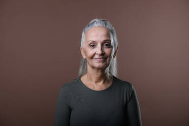 Studio portrait of beautiful senior woman with long gray hair in ponytail. Isolated on a brown pink background. Copy space. - HPIF34661