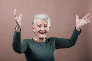 Studio-Porträt einer lächelnden älteren Frau mit erhobenen Armen, kurzen grauen Haaren, grünem Oberteil, isoliert auf braunem, rosa Hintergrund, Kopierraum. - HPIF34664