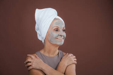 Porträt einer schönen älteren Frau mit Gesichtsmaske und Haarturban Handtuch. Selbstpflege und Haut Routine Konzept. Isoliert auf einem braunen rosa Hintergrund. Kopierraum. - HPIF34677