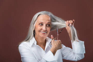 Porträt einer älteren Frau, die ihr schönes graues Haar abschneidet. Haare schneiden wegen Brustkrebs. Konzept zur Sensibilisierung für Brustkrebs. - HPIF34686