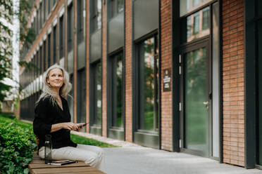 Portrait of beautiful mature woman in middle age with long gray hair, sitting on the bench in the city. - HPIF34706