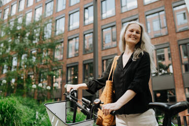Eine schöne Frau mittleren Alters, die mit dem Fahrrad durch die Stadt pendelt, steht neben einem Fahrradständer und ihrem Fahrrad. Eine weibliche Stadtpendlerin, die nach einem langen Arbeitstag mit dem Fahrrad von der Arbeit kommt. - HPIF34715
