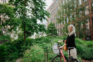 A beautiful middle-aged woman, who commutes through the city by bike, standing next to a bike rack and her bicycle. Female city commuter traveling from work by bike after a long workday. - HPIF34716