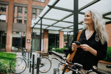 A beautiful middle-aged woman, who commutes through the city by bike, standing next to a bike rack and her bicycle. Female city commuter traveling from work by bike after a long workday. - HPIF34717