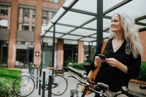 Eine schöne Frau mittleren Alters, die mit dem Fahrrad durch die Stadt pendelt, steht neben einem Fahrradständer und ihrem Fahrrad. Eine weibliche Stadtpendlerin, die nach einem langen Arbeitstag mit dem Fahrrad von der Arbeit kommt. - HPIF34717