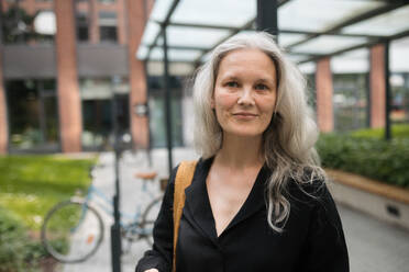 Portrait of middle-aged woman, who commutes through the city by bike, standing next to a bike rack and her bicycle. Female city commuter traveling from work by bike after a long workday. Renting city bike using mobile app. - HPIF34719