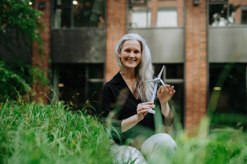 Porträt einer schönen reifen Frau mittleren Alters mit langen grauen Haaren, die ein Modell einer Windturbine hält. Konzept der erneuerbaren Ressourcen. Bedeutung alternativer Energiequellen und langfristige Nachhaltigkeit. - HPIF34728