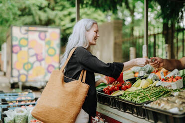 Porträt einer schönen reifen Frau, die auf einem Markt in der Stadt einkauft. Frau mittleren Alters kauft frisches Gemüse und Obst an einem Marktstand. - HPIF34733