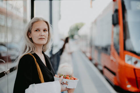 Porträt einer schönen Frau mittleren Alters mit grauen Haaren, die an einer Bushaltestelle auf den Bus wartet. Weiblicher Stadtpendler, der nach einem langen Arbeitstag mit dem Bus von der Arbeit kommt. - HPIF34746