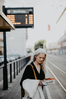 Portrait of a beautiful woman in middle age with gray hair, waiting for the bus at a bus stop. Female city commuter traveling from work by bus after a long workday. - HPIF34747