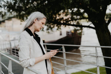 Portrait of beautiful mature woman in middle age with long gray hair, in city using her smartphone. - HPIF34753