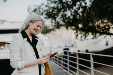 Portrait of beautiful mature woman in middle age with long gray hair, in city using her smartphone. - HPIF34754