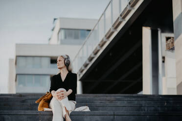 Porträt einer schönen Frau mit grauen Haaren, die auf einer Betontreppe in der Stadt sitzt und über drahtlose Kopfhörer Musik hört. Banner mit Kopierbereich. - HPIF34763