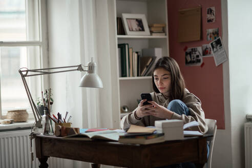 Junges Teenager-Mädchen macht Hausaufgaben und scrollt auf ihrem Smartphone in ihrem Zimmer. - HPIF34782