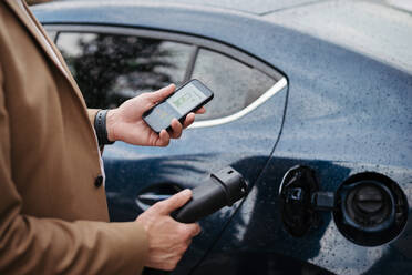 Nahaufnahme eines Geschäftsmannes, der eine App zum Aufladen von Elektrofahrzeugen nutzt, um den Ladevorgang seines Elektroautos über sein Smartphone zu überprüfen. Lade-Apps zur Überwachung des Stromverbrauchs und zum Auffinden von Ladestationen. - HPIF34831
