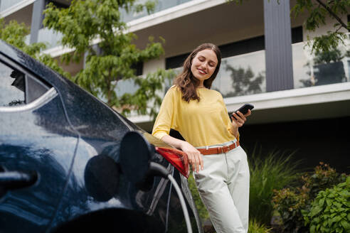 Smiling woman plugging charger in her electric car. Young businesswoman in front house waiting to charge electric vehicle. Female driver using electric vehicle charging app, checking progress of charging of electric car on smart phone. - HPIF34841