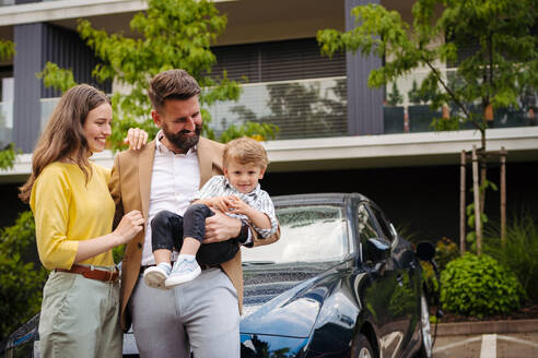 Glückliche Familie steht vor ihrem Elektroauto und lädt es auf der Straße auf. Elektrofahrzeug mit Ladegerät im Ladeanschluss. - HPIF34845