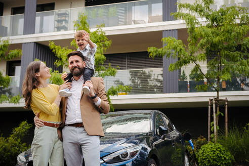 Glückliche Familie steht vor ihrem Elektroauto und lädt es auf der Straße auf. Elektroauto mit Ladegerät im Ladeanschluss. Vater trägt Sohn auf den Schultern. - HPIF34846