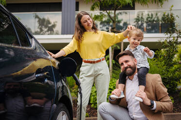 Glückliche Familie, die neben ihrem Elektroauto steht und es auf der Straße auflädt. Elektrofahrzeug mit Ladegerät im Ladeanschluss. Vater hält seinen Sohn auf den Schultern. - HPIF34849