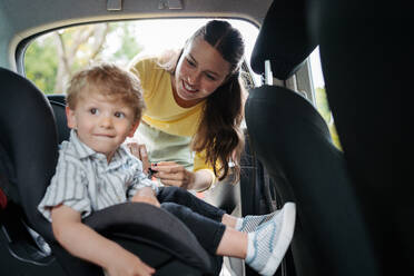 Junge Mutter, die ihren kleinen Sohn in den Autositz setzt und das Kleinkind richtig im Kindersitz anschnallt. - HPIF34851