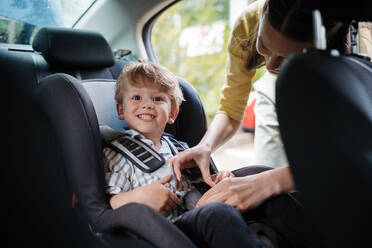 Junge Mutter, die ihren kleinen Sohn in den Autositz setzt und das Kleinkind richtig im Kindersitz anschnallt. - HPIF34852