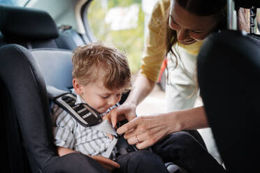 Junge Mutter, die ihren kleinen Sohn in den Autositz setzt und das Kleinkind richtig im Kindersitz anschnallt. - HPIF34853