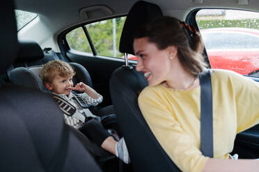 Niedlicher kleiner Junge sitzt in seinem Autositz und spielt auf einem Tablet. Mutter überprüft ihren Sohn vor der Fahrt im Auto. Ride-on Auto Sicherheit. Fahren im Regen. - HPIF34854