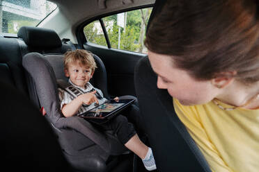 Süßer kleiner Junge sitzt in seinem Autositz und spielt auf einem Tablet. Mutter überprüft ihren Sohn vor der Fahrt im Auto. Ride-on Auto Sicherheit. - HPIF34859