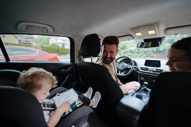 Niedlicher kleiner Junge sitzt in seinem Autositz und spielt auf einem Tablet. Eltern überprüfen ihren Sohn vor der Fahrt im Auto. Sicherheit im Auto. - HPIF34863