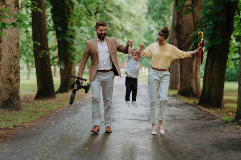 Rückansicht einer jungen Familie mit kleinem Sohn bei einem Spaziergang in einem Stadtpark nach der Arbeit. Berufstätige Eltern verbringen Zeit mit ihrem Sohn nach dem Arbeitstag. Die Eltern holen ihr Kind von der Kindertagesstätte ab. - HPIF34877