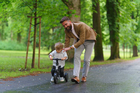 Alleinerziehender Vater, der seinem kleinen Sohn in einem öffentlichen Park das Fahrradfahren beibringt. Berufstätige Eltern, die nach der Arbeit Zeit mit ihrem Sohn verbringen. - HPIF34880