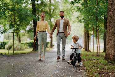 Junge Familie mit kleinem Sohn auf einem Spaziergang in einem Stadtpark nach der Arbeit. Berufstätige Eltern verbringen Zeit mit ihrem Sohn nach dem Arbeitstag. Eltern holen ihr Kind von der Kindertagesstätte ab. - HPIF34881