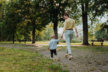 Rückansicht einer Mutter mit ihrem kleinen Sohn bei einem Spaziergang nach der Arbeit. Eine berufstätige Mutter holt ihren Sohn von der Kindertagesstätte ab. - HPIF34889