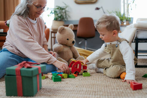 Großmutter spielt mit ihrem kleinen Enkel und Teddybär, der mit einem roten Feuerwehrauto aus Holz spielt. - HPIF34948