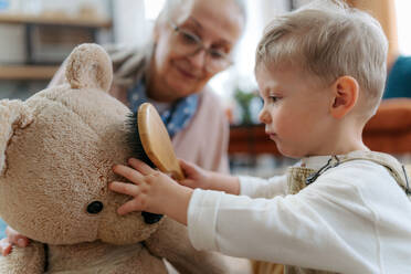 Little boy and grandmother take care of the stuffed toy. Child combing teddy bear's fur. - HPIF34951