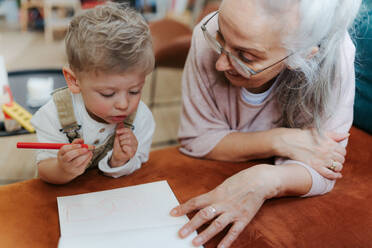Enkel, der mit seiner Großmutter in einen Notizblock zeichnet. Kleiner Junge, der mit Buntstiften zeichnet. - HPIF34995