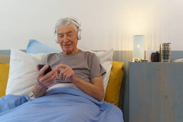 Ein älterer Mann liegt in einem Bett und genießt Musik über Kopfhörer. - HPIF35020