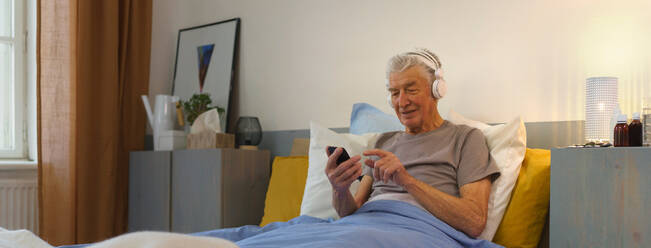 Ein älterer Mann liegt in einem Bett und genießt Musik über Kopfhörer. - HPIF35063