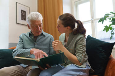 Grandfather with his granddaughter looking at family photo album. Nostalgic senior man showing photos of himself from his youth, remebering old days through pictures. - HPIF35073